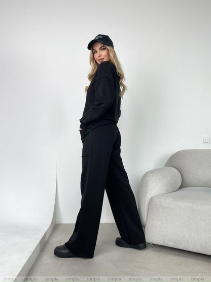 Person wearing a black outfit with a cap in a minimalistic room.