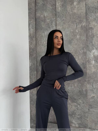 Woman wearing a dark gray long-sleeve top and pants against a gray stone wall.