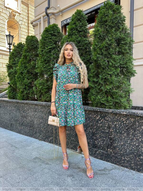 Woman in a floral dress standing outdoors with greenery and a building in the background