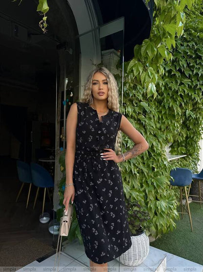 Woman in a black floral dress standing outdoors with greenery and chairs in the background