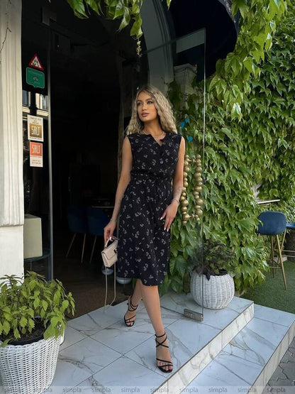 Woman in a dark floral dress standing outdoors with greenery and a building entrance in the background.