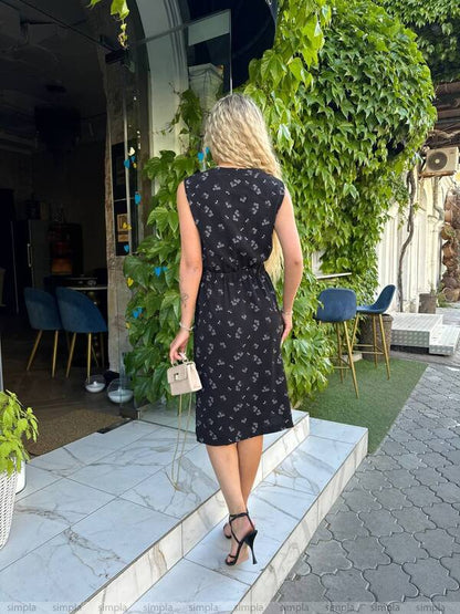 Woman in a black patterned dress standing outdoors near greenery and chairs.