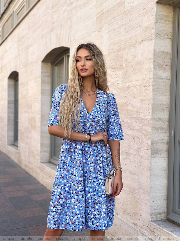Woman wearing a blue floral dress standing against a light-colored wall.