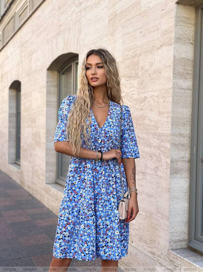 Woman wearing a blue floral dress standing against a light-colored wall.