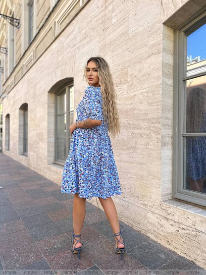Woman in a blue floral dress standing against a light-colored building.
