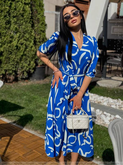 Woman in a blue patterned dress standing outdoors with a white handbag.