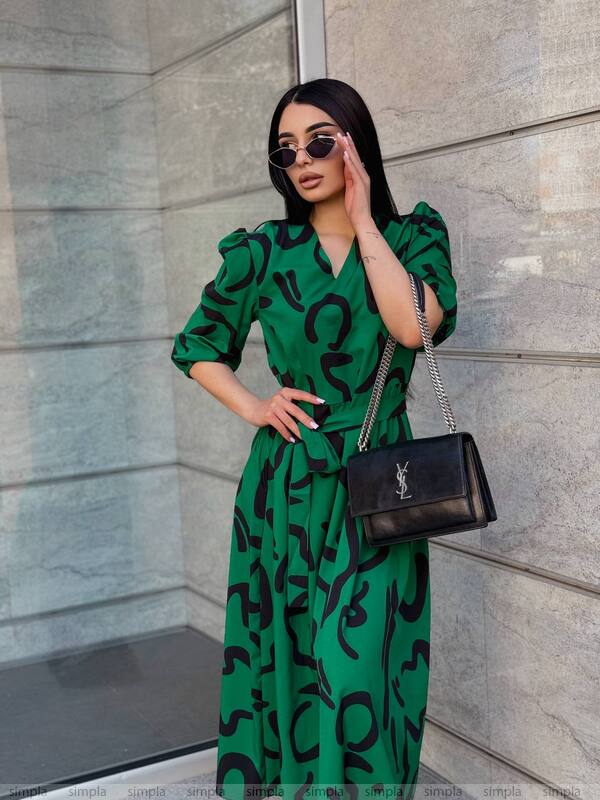 Woman in a green dress with black patterns, holding a black handbag against a gray wall.