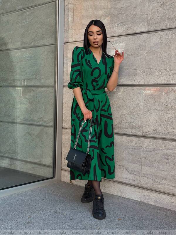 Woman in a green dress with black patterns standing against a gray wall.