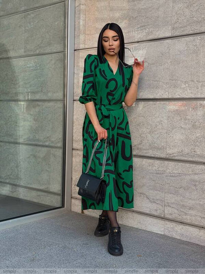 Woman in a green dress with black patterns standing against a gray wall.