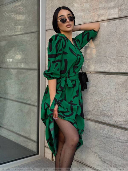 Woman in a green dress with black patterns leaning against a gray wall.