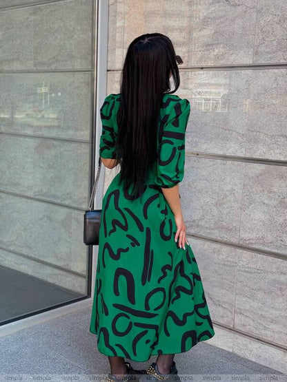 Person wearing a green dress with black patterns standing against a gray wall.