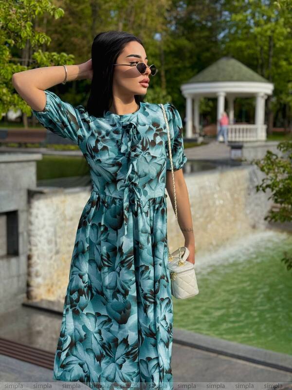 Woman in a blue floral dress standing in a park with a gazebo and pond in the background.
