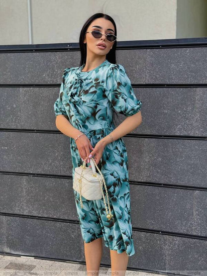 Woman wearing a blue floral dress holding a white handbag against a gray wall.