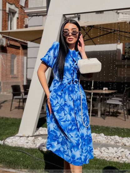 Woman in a blue floral dress standing outdoors with a building and chairs in the background