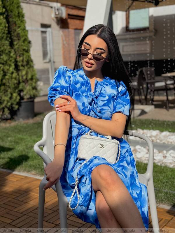 Woman in a blue floral dress sitting outdoors with a white handbag.