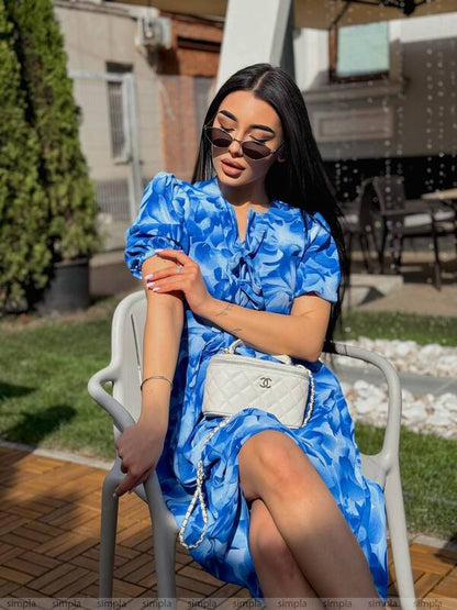 Woman in a blue floral dress sitting outdoors with a white handbag.
