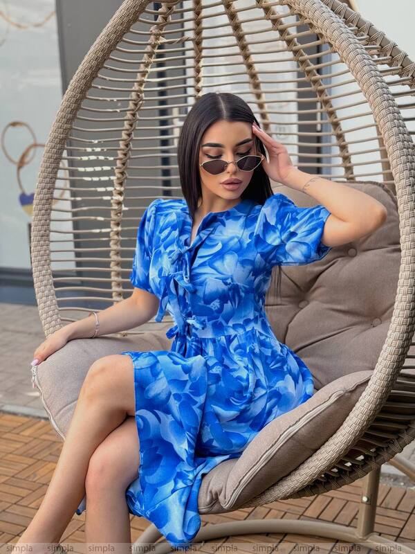 Woman in a blue floral dress sitting in a wicker chair outdoors.