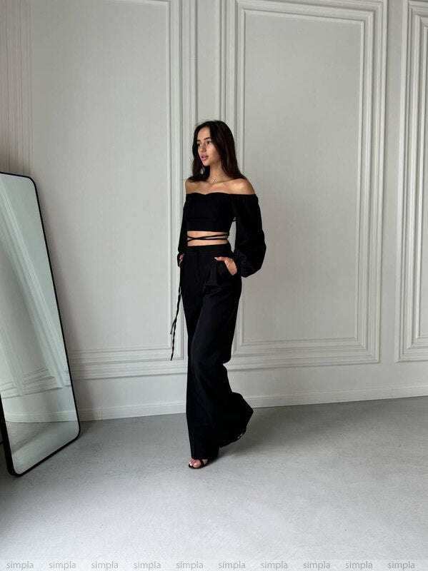 Woman in a black outfit standing in a minimalistic room with white walls and a large mirror.