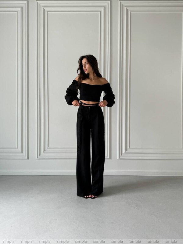 Woman wearing a black off-shoulder top and wide-leg pants against a white paneled wall.
