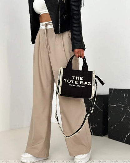 Person holding a black tote bag with 'The Tote Bag' text, wearing beige pants and a black jacket.