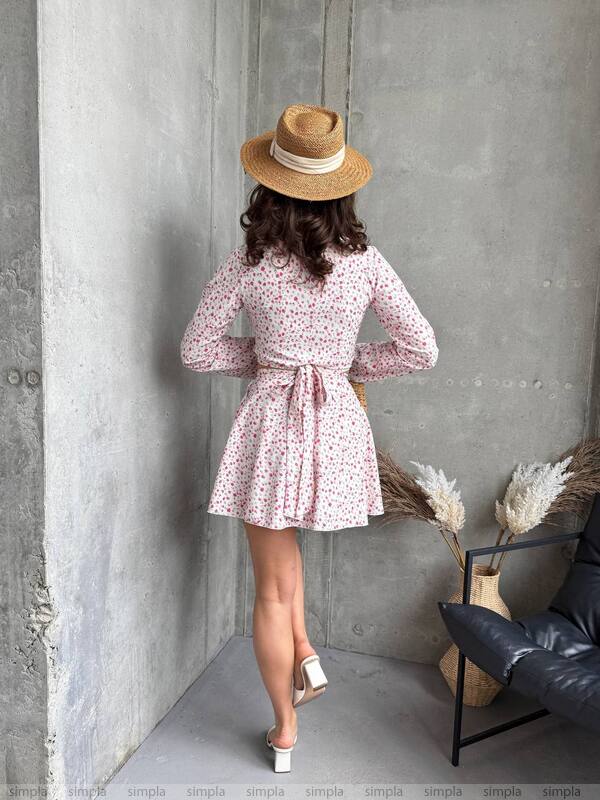 Woman in a floral dress and straw hat standing against a gray wall.
