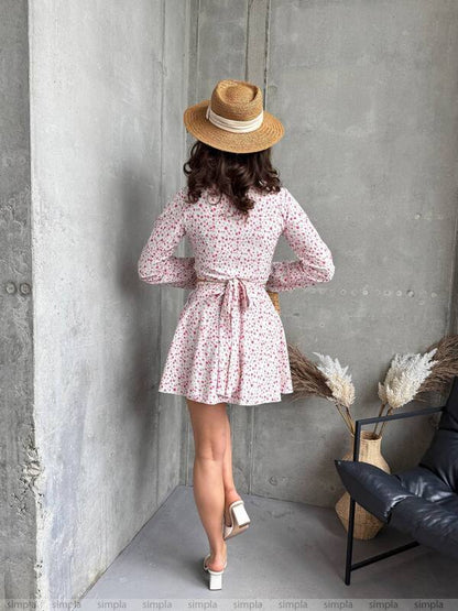 Woman in a floral dress and straw hat standing against a gray wall.