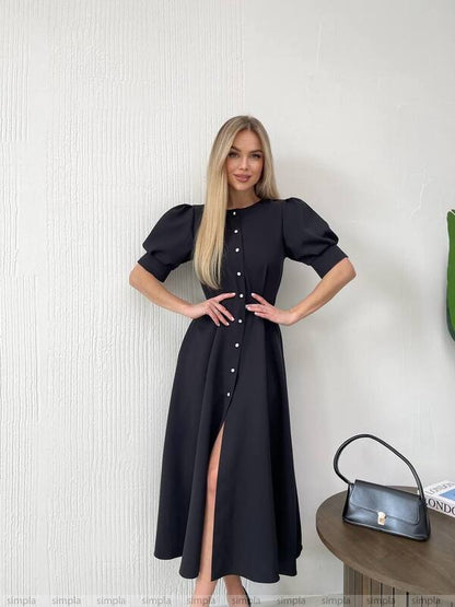 Woman wearing a black dress with a high slit, standing against a light-colored wall.
