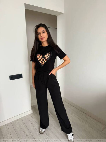 Woman wearing a black t-shirt with a leopard print heart design and black pants in a minimalistic room.