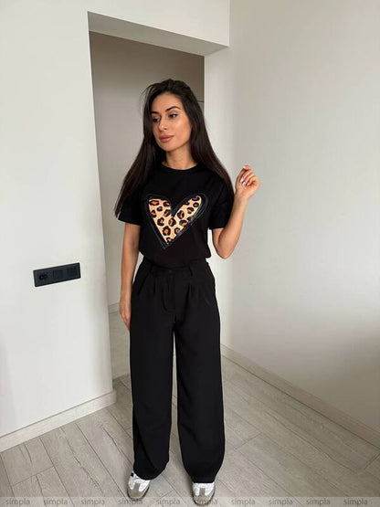 Woman wearing a black t-shirt with a leopard print heart design and black pants in a minimalistic room.