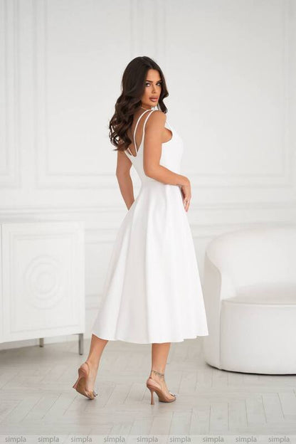 Woman wearing a white dress standing in a minimalistic room with a white chair.