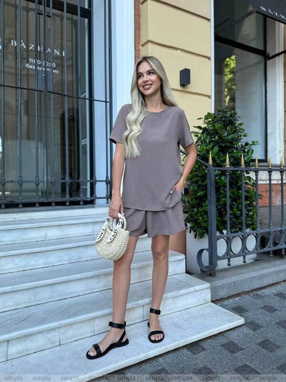 Woman in a gray outfit standing outside a building with plants and steps.