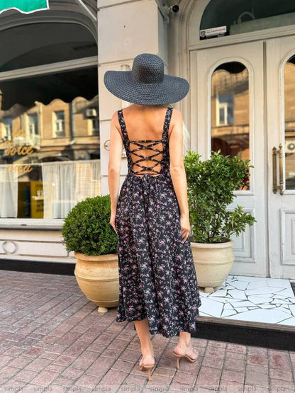 Woman in a floral dress and wide-brimmed hat standing on a street with storefronts and plants.