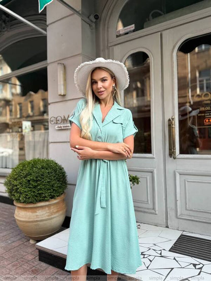 Woman in a light blue dress and white hat standing in front of a building entrance.