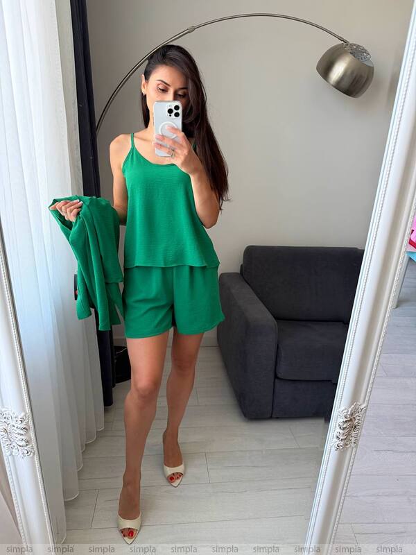Woman taking a mirror selfie wearing a green outfit in a room with a gray sofa and white walls.