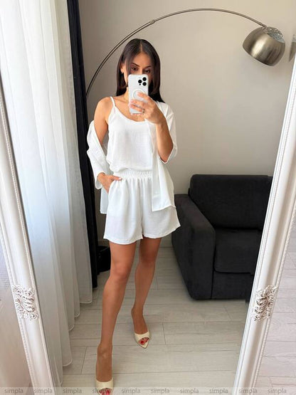 Woman taking a mirror selfie wearing a white outfit in a room with a gray sofa and white curtains.