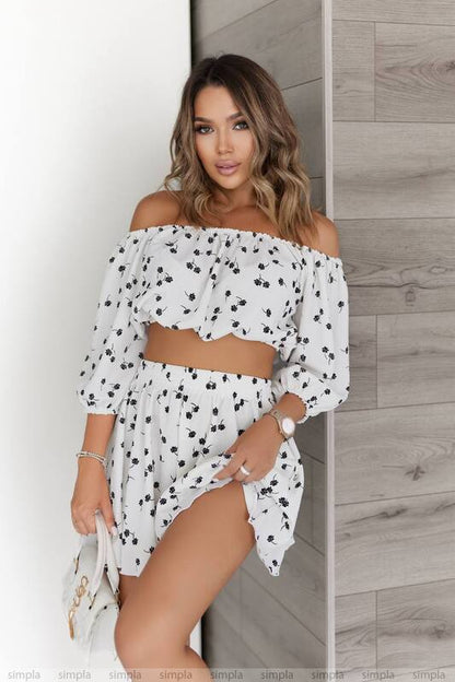 Woman wearing a white off-shoulder outfit with black polka dots against a wooden wall.