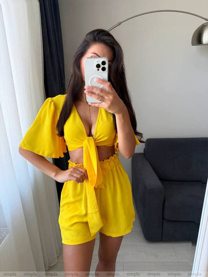 Woman taking a mirror selfie wearing a yellow outfit in a room with a gray sofa and white curtains.