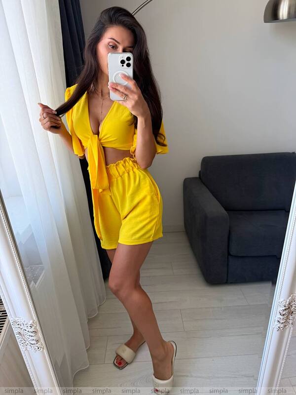 Woman in a yellow outfit taking a mirror selfie in a room with a gray sofa.