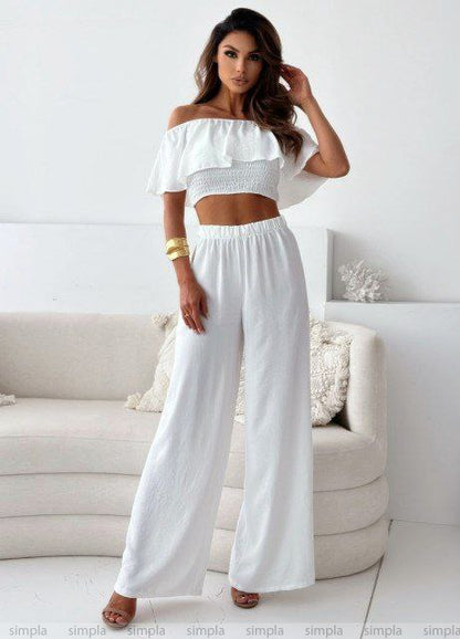 Woman wearing a white off-shoulder top and wide-leg pants in a minimalistic room.