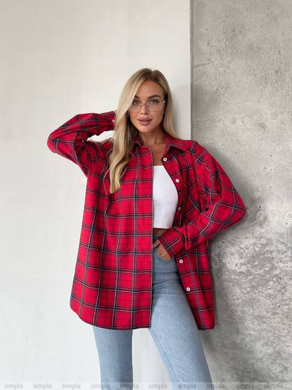 Woman wearing a red plaid shirt over a white top and light blue jeans against a gray wall.