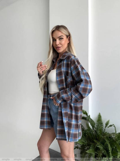 Woman wearing a plaid shirt over a white top and denim shorts, standing against a plain wall with a plant in the background.