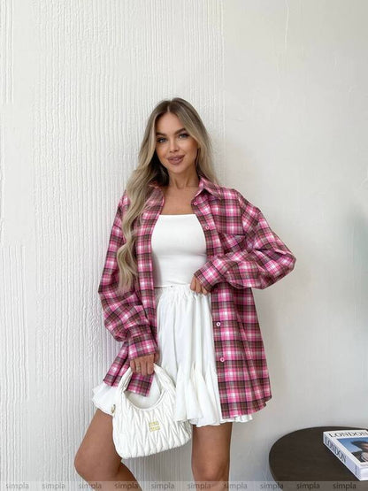 Woman wearing a pink plaid shirt over a white dress against a white wall.