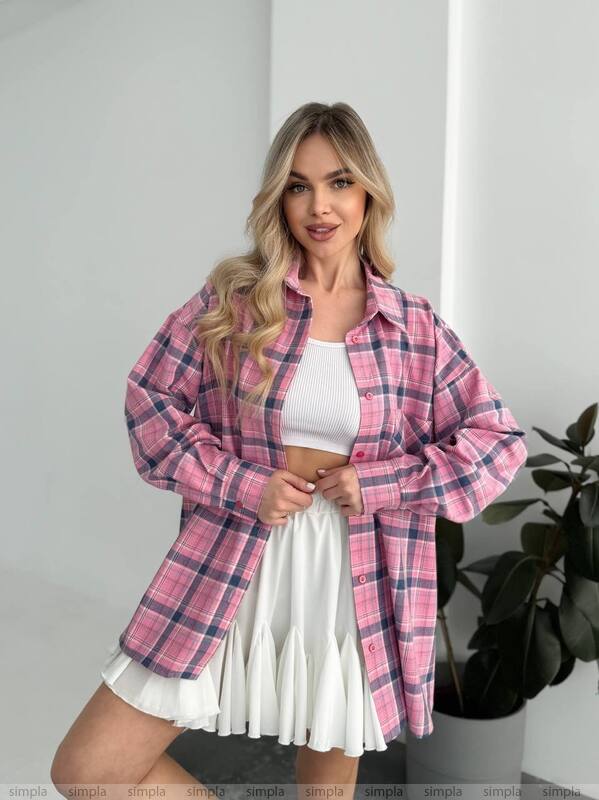 Woman wearing a pink plaid shirt over a white top and skirt in a minimal indoor setting.