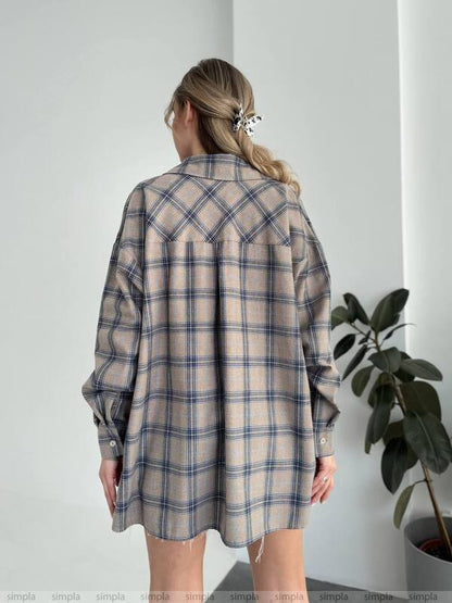 Person wearing a plaid shirt over a dress against a white wall with a plant on the right.