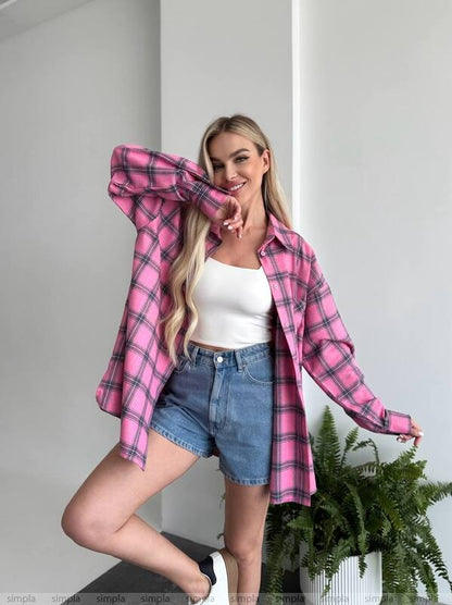Woman wearing a pink plaid shirt over a white top and denim shorts, standing indoors with a plant in the background.