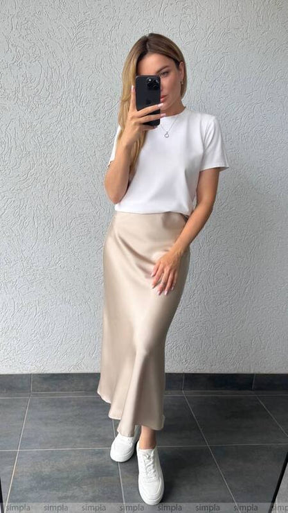 Person wearing a white t-shirt and beige skirt against a plain wall.