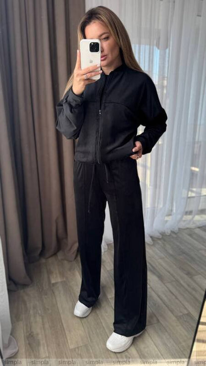 Person wearing a black outfit taking a mirror selfie in a room with curtains.