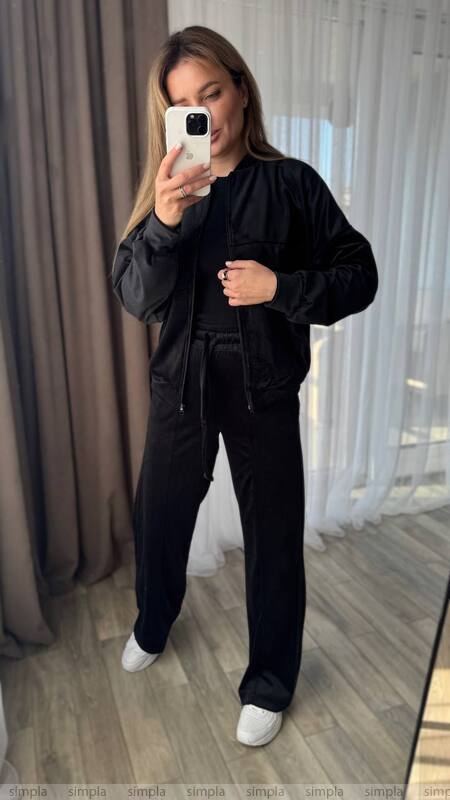 Person wearing a black outfit taking a mirror selfie in a room with curtains.
