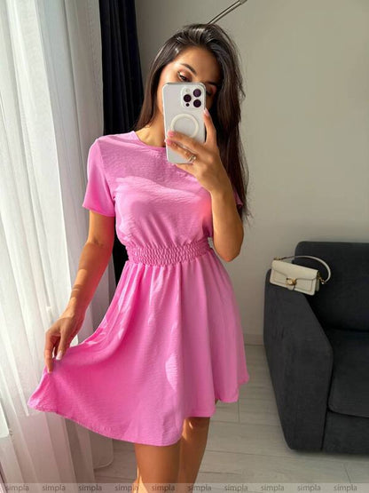 Woman wearing a pink dress taking a mirror selfie in a room with a couch and white curtains.