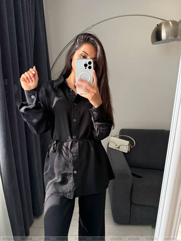 Person wearing a black jacket and pants taking a mirror selfie in a room with gray curtains and a sofa.